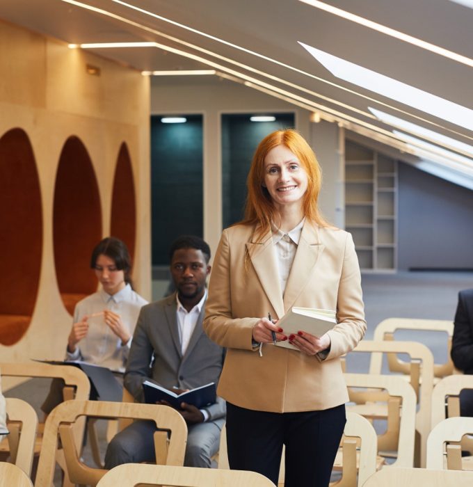 Portrait of mature red-haired businesswoman smiling cheerfully looking at camera while standing among audience in office or conference hall, copy space